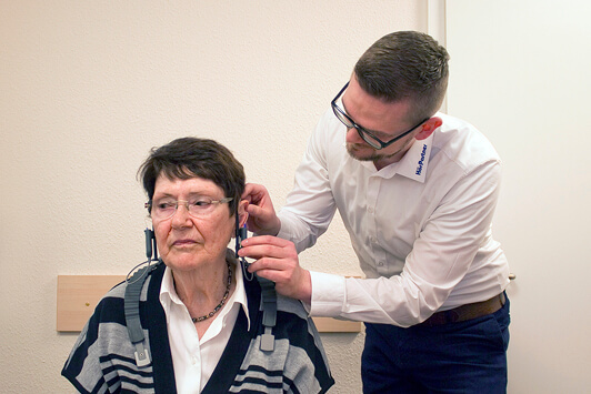 Ein Hörakustiker passt einer älteren Frau ein Hörgerät im Beratungsraum an.